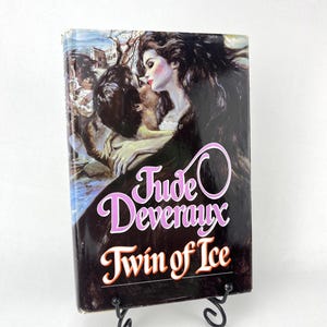 Può includere: Un libro tascabile con una copertina nera e un dipinto di una coppia che si bacia. Il titolo del libro è "Jude Deveraux Twin of Ice".