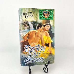May include: A paperback book titled "Tempting Texas Treasure" by Wanda Owen, displayed on a black metal stand. The cover art depicts a couple embracing near a waterfall. The woman wears a yellow dress.