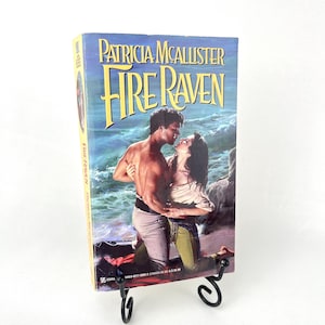 May include: A paperback novel titled "Fire Raven" by Patricia McAllister. The cover features a couple embracing on a beach with a dramatic ocean backdrop.