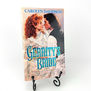 May include: A paperback book titled "Gerrity's Bride" by Carolyn Davidson, displayed on a black metal stand. The cover features a romantic illustration of a man and woman. The book is part of the Harlequin Historicals series.