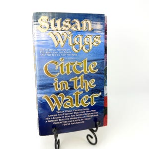 以下が含まれることがあります： スーザン・ウィッグス著のペーパーバック『Circle in the Water』。青い水面の背景に金色のエンボス加工された文字が特徴です。表紙には著者に関するテキストとジェイン・アン・クレンツによる引用文が記載されており、黒い金属製のスタンドに立てかけられています。
