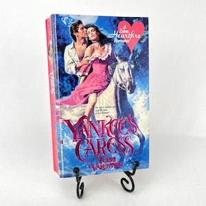 May include: A paperback book titled "Yankee's Caress" by Terri Valentine. The cover features a romantic illustration of a woman in a pink dress on a white horse, embraced by a man. The book is displayed on a black metal stand.