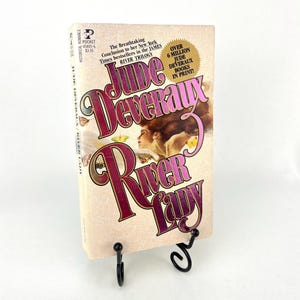 Può includere: Un libro tascabile intitolato "River Lady" di Jude Deveraux, esposto su un supporto in metallo nero. La copertina presenta un ritratto di donna e il titolo in un carattere decorativo. Il libro fa parte della trilogia James River.