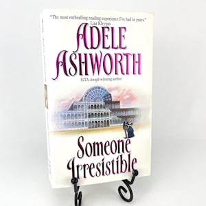 Puede incluir: Un libro de bolsillo titulado "Someone Irresistible" de Adele Ashworth, con una cita de Lisa Kleypas. La portada presenta una ilustración de un edificio y una pareja. El libro se exhibe en un soporte de metal negro.