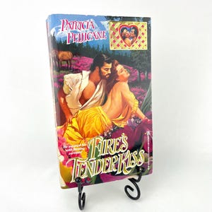 May include: A paperback romance novel with a pink, purple, and green cover featuring a couple embracing in a field of flowers. The title of the book is "Fire's Tender Kiss" by Patricia Pelican. The book is on a black metal book stand.