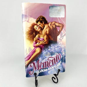 May include: A paperback book titled "Memento" by Leanora Brownleigh, displayed on a black metal stand. The cover art features a romantic illustration of a man and woman embracing, set against a purple and blue background.