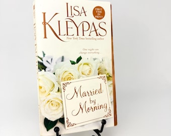 Op de afbeelding: Een paperback getiteld Married by Morning van Lisa Kleypas, met een omslag met witte rozen en de tekst First Time in Print en One night can change everything. Het boek staat rechtop op een zwarte metalen draadstandaard.