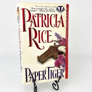 May include: A paperback book titled "Paper Tiger" by Patricia Rice. The cover features a vintage-style gun and flowers in shades of pink and purple. The book is on a black metal book stand.