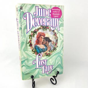 Può includere: Un libro tascabile intitolato "Lost Lady" di Jude Deveraux, autrice di best seller di Mountain Laurel. La copertina presenta una coppia che si abbraccia in una ghirlanda di fiori.