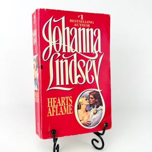 Pode incluir: Um livro de capa dura vermelho intitulado "Hearts Aflame" de Johanna Lindsey. A capa apresenta uma ilustração romântica de um casal e o texto "#1 Bestselling Author". O livro é exibido em um suporte de metal preto.