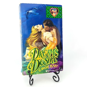 May include: A paperback book titled "Dreams Desire" by Gwen Cleary. The cover features a romantic illustration of a man and woman embracing, with a blue background and a seagull. The book is displayed on a black metal stand.