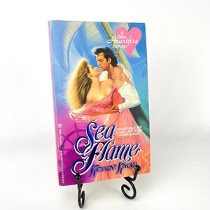 May include: A paperback book titled "Sea Flame" by Katharine Kincaid, displayed on a black metal stand. The cover features a romantic illustration of a couple embracing, with the words "A Zebra Heartfire Romance" at the top.