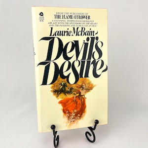 Pode incluir: Um livro de bolso vintage intitulado "Devil's Desire" de Laurie McBain. A capa apresenta uma mulher com um vestido vermelho correndo por um campo com cavalos ao fundo. O livro é dos editores de "The Flame and the Flower".