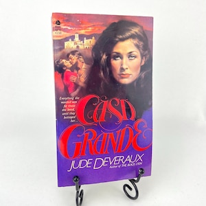 Può includere: Un libro tascabile intitolato "Casa Grande" di Jude Deveraux. La copertina presenta un ritratto di donna, una coppia che si abbraccia e una scena di tramonto. Il libro è esposto su un supporto di metallo nero.