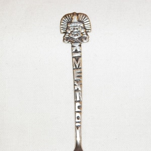 May include: Silver souvenir spoon with a detailed design of a Mayan head and the word "MEXICO" engraved on the handle.