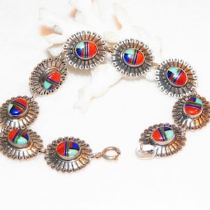 Op de afbeelding: Een zilveren armband met zeven ovale turquoise, koraal en lapis lazuli inlegstenen. Elke steen is omgeven door een zilveren zonnestralenontwerp.