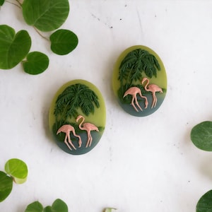 May include: Two green and blue oval-shaped cabochons with a raised design of pink flamingos and palm trees.