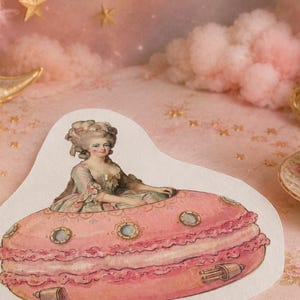 May include: A die-cut sticker featuring an illustration of a person with an elaborate 18th-century hairstyle and dress sitting inside a large, pink, circular macaron shaped like a flying saucer with small portholes and metallic thrusters.