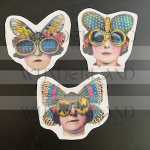 May include: Three vintage-style paper cutouts of women wearing butterfly-shaped goggles. Each cutout features a different butterfly design with colorful wings and intricate details. The goggles are round and have a steampunk aesthetic.