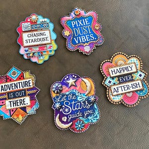 May include: Five colorful, round, decorative magnets with inspirational quotes. The magnets feature a variety of designs, including stars, moons, and flowers. The quotes on the magnets are: "Chasing Stardust", "Pixie Dust Vibes", "Adventure is Out There", "Let the Stars Guide You", and "Happily Ever After-ish".