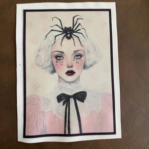 May include: A white canvas patch with a black spider on top of a woman's head. The woman has white hair, pink cheeks, and is wearing a pink dress with a black bow.