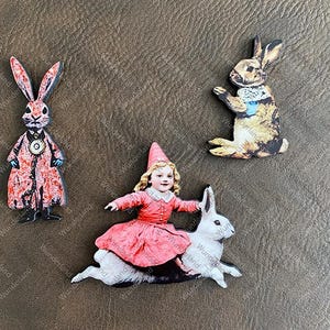 May include: Three wooden ornaments featuring whimsical illustrations of rabbits. One rabbit is wearing a red coat and a pocket watch, another is brown and white with a blue bow, and the third is white with a girl in a red dress riding on its back.