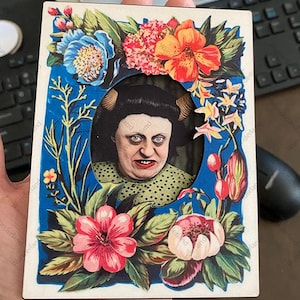 May include: A rectangular card featuring a vintage portrait of a woman with horns, framed by a blue border and colorful floral arrangements. The woman has a stern expression. The flowers include blue, orange, pink, and red blooms.