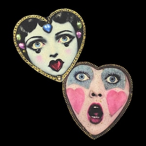 May include: Two gold heart-shaped frames with images of women's faces. The top frame features a woman with blue eyes, red lips, and a black wig. The bottom frame features a woman with blue eyes, a surprised expression, and pink cheeks.