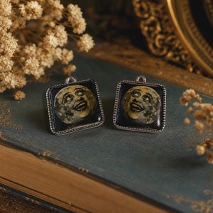 May include: Two square pendants featuring a vintage-style illustration of a smiling moon with a textured, cratered surface. Each pendant is encased in a silver-toned metal frame with a small loop at the top for attachment to a chain or cord.