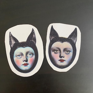 May include: Two sticker decals featuring a vintage illustration of a face with blue eyes, red lips, and rosy cheeks. The face is framed by a black cap with pointed ears and a starry pattern. The stickers are on a white background.