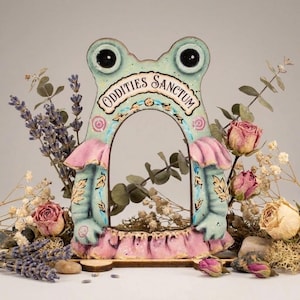 May include: A whimsical frog-shaped decorative frame in pastel colors. The frame features the text "ODDITIES SANCTUM" and is adorned with dried flowers and lavender. The frog has large, round eyes and a pink and blue color scheme.