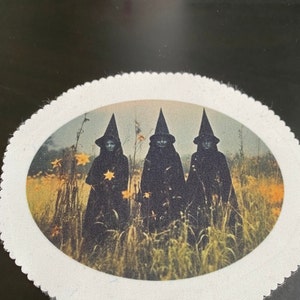 May include: An oval-shaped patch featuring a vintage image of three figures in black cloaks and pointed hats, standing in a field of tall grass and yellow flowers. The figures appear to be witches, with one holding a star. The patch has a white, scalloped border.