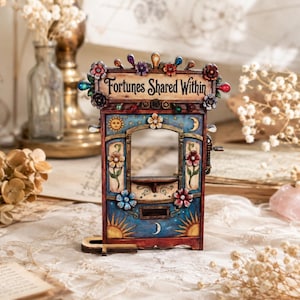 May include: A rectangular wooden fortune teller booth with a blue and red finish, featuring the text "Fortunes Shared Within" at the top. The structure is decorated with painted suns, moons, and floral motifs, with small colorful accents along the top edge.