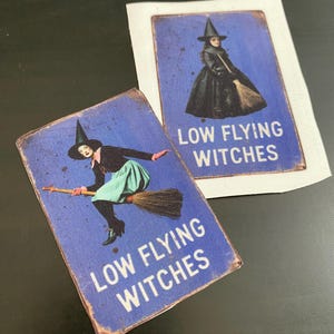 May include: Two blue and white signs with a witch on a broomstick and the text "Low Flying Witches".