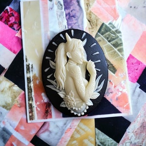 May include: An oval-shaped cameo pendant featuring a cream-colored, detailed relief of a female figure with horns, a ponytail, and tattoos. The figure is set against a black background with a white border. The pendant is on a colorful, textured background.