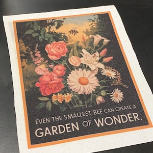 May include: A white canvas patch with a black background featuring a vintage illustration of a bouquet of flowers with a bee flying above. The text "EVEN THE SMALLEST BEE CAN CREATE A GARDEN OF WONDER." is printed in white on the bottom of the patch.