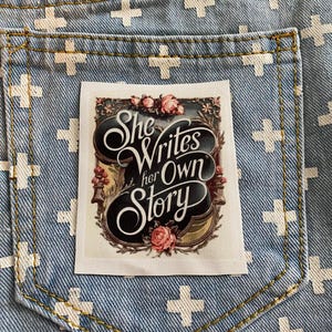May include: A white patch with a floral design and the text "She Writes her Own Story" in black script. The patch is sewn onto a blue denim background with white crosses.