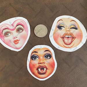 May include: Three colorful stickers featuring different faces. The first sticker is a pink heart with a smiling face. The second sticker is a round face with a wide open mouth and tongue sticking out. The third sticker is a round face with a wide open mouth and fangs.