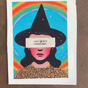 May include: A fabric patch featuring a stylized illustration of a person wearing a black witch hat. The image has a rainbow and blue background with the text "NOT QUITE ORDINARY."
