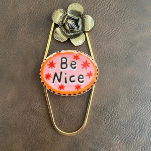 May include: A gold-colored metal bookmark with a flower and an oval pendant. The pendant has the words "Be Nice" in black letters on a pink background with orange trim and red star accents. The bookmark is on a brown surface.