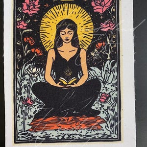 May include: A tarot card with a black background and a woman sitting in the center. The woman is wearing a black dress and has a halo of light around her head. She is holding a book in her lap. The card is surrounded by flowers and stars.