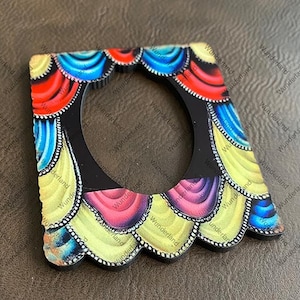 May include: A colorful, square-shaped decorative frame with a circular opening. The frame features a black base with a scalloped edge and is adorned with vibrant hues of red, blue, yellow, and pink.