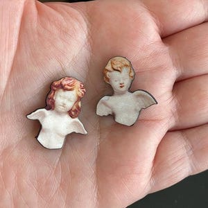 May include: Two small, wooden angel-shaped earrings. Each earring features a cherubic face with closed eyes, small red lips, and light brown hair. The angels have white bodies and small wings. The earrings are set against a light skin tone.