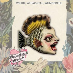 May include: A whimsical enamel pin featuring a stylized fish with a human face and yellow hair. The fish is green, gray, and white with red and black accents. The pin is set against a background of floral illustrations. The text "Wonderland Woodcuts" is visible in a pink heart shape.