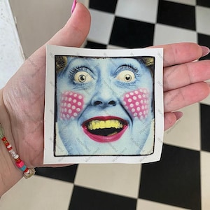 May include: A square sticker featuring a blue face with wide eyes, a red mouth, and yellow teeth. The cheeks have pink polka dots. The sticker is held in a hand against a black and white checkered background.