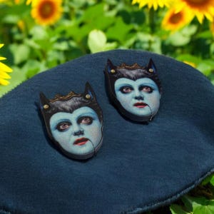 May include: Two face-shaped brooches with blue skin, red lips, and black hair, adorned with a gold crown and cat-like ears. The brooches rest on a dark blue surface, set against a backdrop of sunflowers.