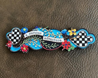 May include: A whimsical, decorative plaque with the words "a curious adventure." The plaque features a blue background with floral and checkered accents in black, white, red, and yellow. The design is set against a brown background.