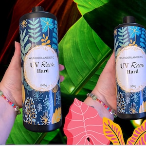 May include: Two bottles of UV Resin Hard, one labeled 1000g and the other 500g. The bottles have a dark blue floral design with a white oval label that reads "UV Resin Hard". The background is a mix of green and red.