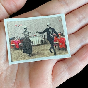 May include: A fabric patch with a vintage-style image of two skeletons dancing in a dining room. The skeletons are dressed in formal attire, and the room is decorated with red and white tablecloths and chairs.