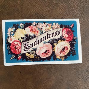 May include: A rectangular patch with a teal border features a floral design. The design includes pink, red, and yellow flowers surrounding a banner with the word "Enchantress" in a decorative font. The patch is likely made of fabric.